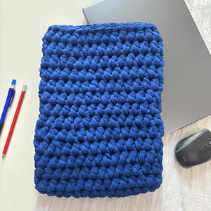Könnte beinhalten: Eine leuchtend blaue, handgehäkelte Laptoptasche. Die Tasche ist rechteckig und aus dickem, strukturiertem Garn gefertigt. Ein silberner Laptop, eine Computermaus, ein blauer Stift und ein roter Bleistift sind ebenfalls zu sehen.