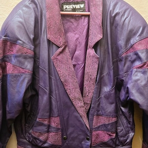 May include: A purple leather jacket featuring a wide collar and panels with a textured snakeskin pattern. The jacket has a double-breasted front with two visible buttons and a label on the inside neck that reads PREVIEW LEATHERS.