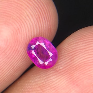 May include: A vibrant, oval-cut gemstone with a deep magenta-pink hue. The stone features a faceted surface that creates a distinct star-like pattern in the center, reflecting light with high clarity and a polished, smooth finish throughout.