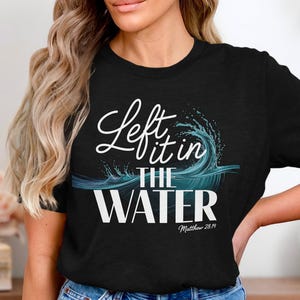May include: A black short-sleeved t-shirt featuring a graphic of a teal and white ocean wave. The text reads Left it in THE WATER with the scripture reference Matthew 28:19 printed in a smaller white font underneath the main design.