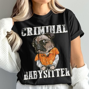 Könnte beinhalten: Ein schwarzes T-Shirt mit einer Grafik eines Mopses, der eine Sonnenbrille und einen orangefarbenen Gefängnisanzug mit Handschellen trägt. Der Text "CRIMINAL" steht in weißer, abgenutzter Schrift darüber und "BABYSITTER" darunter.