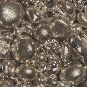 May include: Close-up of a collection of shiny, silver-colored metal beads. The beads vary in size and shape, with a reflective surface that catches the light. The overall effect is a textured, metallic surface.