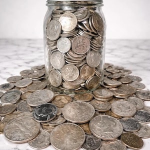 May include: A clear glass jar filled with silver coins, with a silver lid. The jar sits on a marble surface, surrounded by more silver coins. The coins vary in size and design, reflecting different historical periods.