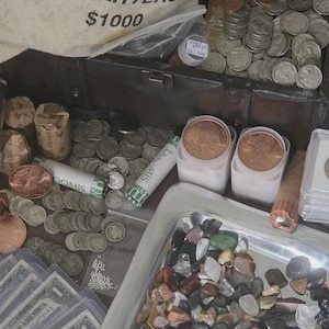 May include: A collection of coins, currency, and gemstones. A canvas bag labeled "U.S. MINT QUARTERS $1000" is open, revealing stacks of quarters. Rolls of coins, paper currency, and a tray of colorful gemstones are also visible.