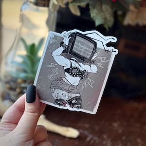 May include: A black and white sticker featuring a muscular figure with a television for a head. The figure is wearing a studded bra, denim shorts, and bracelets. The TV screen displays a heartbeat monitor. Lightning bolt graphics surround the figure.