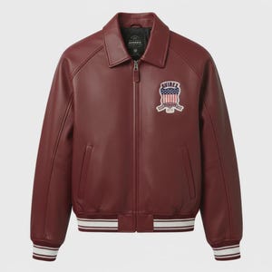Op de afbeelding: Bordeaux leren jas met een kraag en een ritssluiting. De jas heeft een patch op de borst met een patriottisch ontwerp en het woord "AVIREX". De manchetten en zoom hebben wit en zwart gestreepte details.