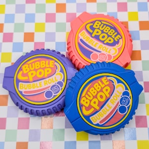 May include: Three colorful Bubble Pop Bubble Roll containers. The containers are in pink, purple, and blue. Each container has a yellow and pink logo with the words "Bubble Pop Bubble Roll". The background is a colorful checkered pattern.
