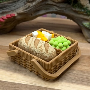 Op de afbeelding: Een miniatuur picknickmand van bruin vlechtwerk, gevuld met een brood, kaas en groene druiven. De mand heeft een handvat en staat op een houten oppervlak.