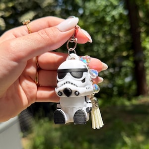 Può includere: Un portachiavi Stormtrooper bianco e nero con chiusura argentata, occhiali da sole neri e nappa color crema. Il portachiavi è adornato da cristalli iridescenti. La figura è una rappresentazione stilizzata di un personaggio del franchise di Star Wars.