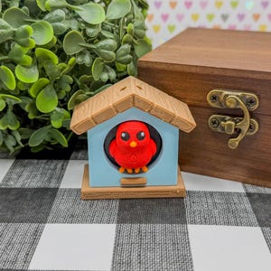 May include: A 3D-printed birdhouse with a red bird inside. The birdhouse is light blue with a brown roof and a small orange perch. A wooden box with a metal clasp is in the background. The scene is set on a black and white checkered surface.