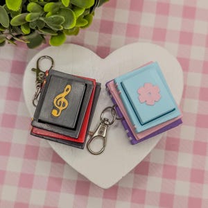 May include: Two miniature book keychains. One is black with a gold treble clef, the other is blue with a pink flower. Both have silver key rings and are on a white heart-shaped surface.