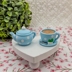 Pode incluir: Um conjunto em miniatura de bule e chávena azul claro com bolinhas brancas. A chávena contém um líquido castanho e assenta num pires com uma colher, um cubo de açúcar e uma folha de hortelã. O conjunto é exibido num suporte branco em forma de coração.
