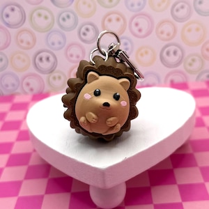 Könnte beinhalten: Ein brauner Igel-Schlüsselanhänger mit silbernem Schlüsselring. Der Igel hat einen hellbraunen Körper, dunkelbraune Stacheln und rosa Wangen. Er steht auf einem weißen herzförmigen Ständer.