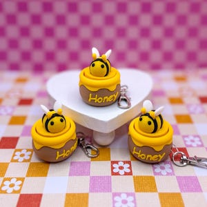 Può includere: Tre portachiavi fatti a mano a tema api. Ogni portachiavi presenta un piccolo vasetto di miele marrone con un coperchio giallo e un'ape nera e gialla sopra. La parola "Honey" è scritta sul vaso. Ogni portachiavi ha un fermaglio argentato.