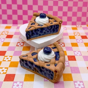 Puede incluir: Dos porciones de tarta de arándanos en miniatura. Cada porción tiene una corteza de rejilla, un relleno de bayas azul oscuro y está cubierta con una cucharada de crema batida blanca y un solo arándano. Las tartas están sobre un soporte blanco en forma de corazón y un fondo estampado colorido.