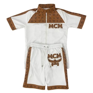 May include: A white and brown two-piece set consisting of a short-sleeved zip-up jacket and matching shorts. The jacket features brown panels with the MCM logo pattern on the shoulders and sleeves. The shorts have a drawstring waist and logo.