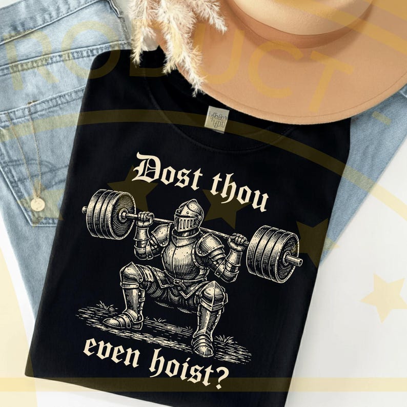 Puede incluir: Camiseta negra con un gr&aacute;fico de un caballero levantando pesas, con el texto "Dost thou even hoist?" en una fuente medieval. El caballero lleva una armadura completa. La camiseta se muestra sobre unos vaqueros azules.