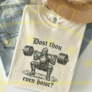 Puede incluir: Camiseta color crema con una ilustraci&oacute;n en blanco y negro de un caballero levantando una barra. El texto "Dost thou even hoist?" est&aacute; impreso encima y debajo del caballero. La camiseta est&aacute; doblada sobre una superficie de mezclilla azul claro.