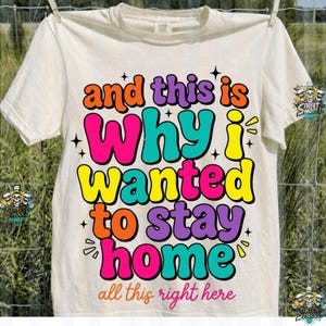 May include: Cream-colored t-shirt with the text "and this is Why I wanted to stay home" in vibrant colors. The text is outlined in black and has a retro, groovy style. The words "all this right here" are at the bottom.