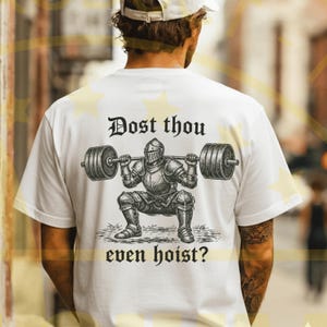 Puede incluir: Camiseta blanca con una ilustraci&oacute;n en blanco y negro de un caballero levantando pesas. El texto "Dost thou even hoist?" est&aacute; en una fuente g&oacute;tica. El dise&ntilde;o muestra a un caballero con armadura completa haciendo una sentadilla con una barra.