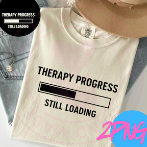 May include: Cream-colored t-shirt with the text "THERAPY PROGRESS" above a loading bar graphic, and "STILL LOADING" below. The shirt is folded, with a Comfort Colors tag visible. A hat and jeans are in the background.