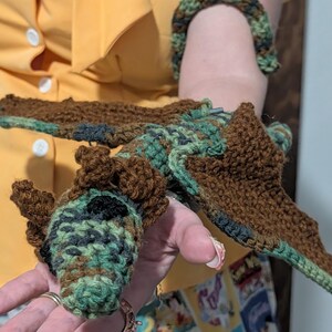May include: A handmade crochet dragon in shades of brown, green, and black. The dragon has wings and a detailed head. A matching crochet bracelet is also visible. The dragon is held in a person's hand.