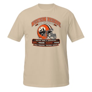 May include: A beige short-sleeved t-shirt featuring a graphic of an orange and brown football helmet with a wolf silhouette. Text includes WESTERN RESERVE LONGHOUSE, NATIVE SONS & DAUGHTERS, MINGO, PENACOOK, BLACKFOOT, KICKAPOO, PALS FOREVER, FRIENDS ALWAYS.