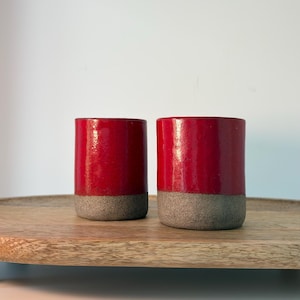 Peut inclure: Deux tasses cylindriques en céramique rouge avec une base grise, posées sur un plateau en bois. Les tasses ont une finition brillante et un design simple. Le plateau présente un veinage naturel.