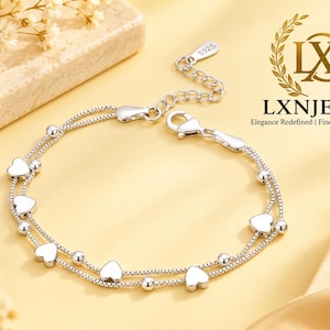 May include: A double-strand silver bracelet featuring small heart-shaped charms and round beads along the chain. The bracelet includes a lobster claw clasp and a small tag stamped with S925. The logo LXN LXNJEWELS is visible in the upper right corner.