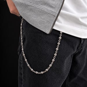 May include: A silver wallet chain featuring a series of repeating circular links with an embossed cross design. The chain is attached to the waistband of dark gray denim jeans, hanging in a curved drape against the fabric of the pants.