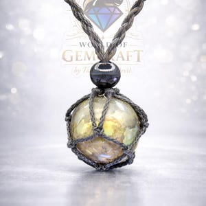 May include: A handcrafted necklace featuring a large, iridescent orb encased in a gray macrame net. The orb is suspended from a gray braided cord, with a black bead accent. The necklace has the text "WORLD OF GEMCRAFT" in the background.