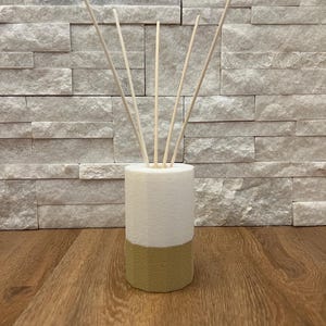 Puede incluir: Un difusor de caña de dos tonos con una sección superior blanca y una base dorada. Varias cañas de madera se insertan en la parte superior. El difusor se encuentra sobre una superficie de madera, con una pared de piedra texturizada en el fondo.