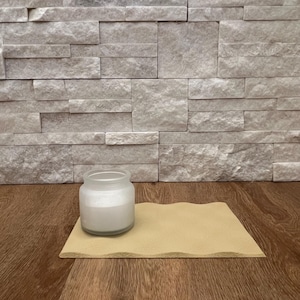 May include: A clear glass jar filled with white wax, sitting on a beige, wavy-edged square. The jar is placed on a wooden surface, with a textured stone wall in the background. The candle is unscented.