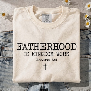 Puede incluir: Una camiseta de color crema con texto negro que dice FATHERHOOD IS KINGDOM WORK, seguido de Proverbs 22:6 y un pequeño símbolo de cruz. La camiseta presenta una etiqueta de la marca Comfort Colors en el cuello interior indicando talla mediana.