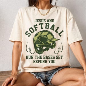 May include: Cream-colored t-shirt with the text "JESUS AND SOFTBALL" above a green softball helmet graphic. Below the helmet, the text reads "RUN THE BASES SET BEFORE YOU". The shirt is worn by a person.