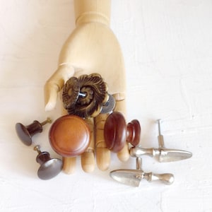 May include: A collection of antique cabinet knobs and pulls in various styles and finishes. The assortment includes dark brown wooden knobs, a decorative bronze knob, and silver-toned metal pulls. The knobs and pulls are displayed on a light background.