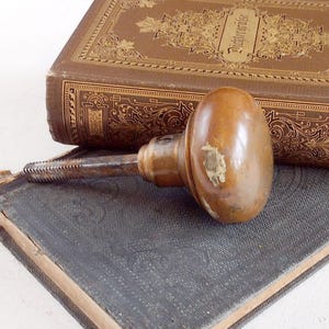 May include: A vintage, oval-shaped, wooden door knob with a metal threaded rod. The knob is a warm brown color with a worn, aged appearance. It rests on a dark blue book with gold embossed lettering and a brown book with gold floral designs.