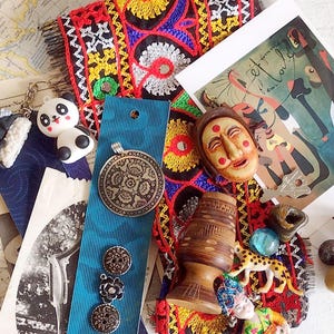 May include: A collection of various decorative items, including a panda bear keychain, a pendant, buttons, a wooden mask, and a small wooden vase. The items are arranged on a colorful embroidered textile and a vintage map.