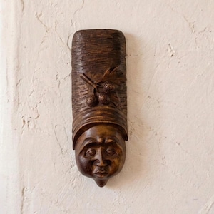 May include: A carved wooden wall hanging featuring a stylized face with a long, rectangular headdress. The dark brown piece has a textured surface and is adorned with carved details, including a face with closed eyes and a small mouth. The top features a carved design of a plant.