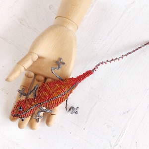 May include: A beaded lizard figurine in shades of red, orange, and yellow, with silver-colored wire legs and a long tail. The lizard is resting on a wooden hand against a white background. The lizard's body is covered in small beads.