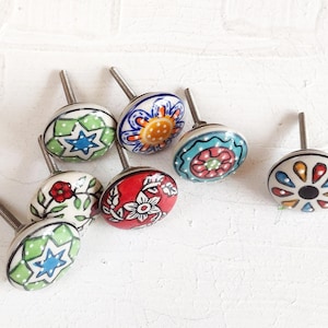 May include: A collection of ceramic cabinet knobs with various hand-painted designs. The knobs feature floral, star, and geometric patterns in colors like red, blue, green, and yellow. Each knob has a silver-colored screw for attachment.