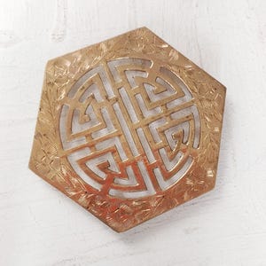 May include: A hexagonal brass trivet featuring an intricate openwork geometric pattern in the center. The outer border is decorated with an engraved floral motif. The metal surface has a warm, polished gold tone with visible etched details.