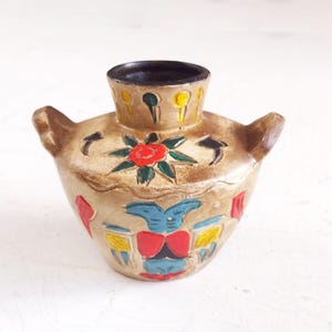 May include: A small, tan ceramic vase with a dark brown interior and two side handles. The vase is decorated with a red flower, blue and yellow geometric shapes, and a floral pattern around the rim. The vase is approximately 4 inches tall.