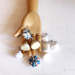 May include: A collection of decorative cabinet knobs in various designs. Some knobs are white with blue floral patterns, others are solid white or cream-colored. One knob features a blue and red patterned design. The knobs are displayed on a wooden hand.