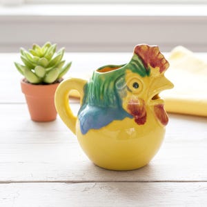 May include: A ceramic pitcher shaped like a rooster with a yellow body, a green neck, and a red comb and wattle. The pitcher features a curved handle on the side and an open top for pouring, with a glossy finish and a rounded base design.