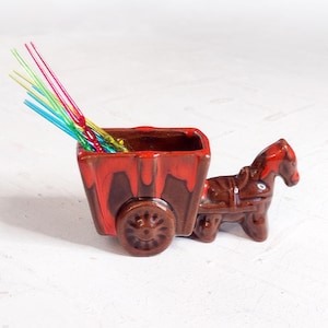 May include: A ceramic horse-drawn cart with a brown and red glaze. The cart holds colorful cocktail stirrers. The horse and cart are detailed with wheels and a rectangular container. A decorative piece for a bar or kitchen.