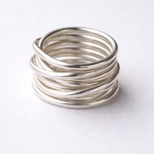 Puede incluir: Un anillo plateado compuesto por múltiples bandas enrolladas. El anillo tiene un acabado liso y brillante. Las bandas están enrolladas firmemente, creando un diseño único y moderno. El anillo está sobre un fondo blanco.