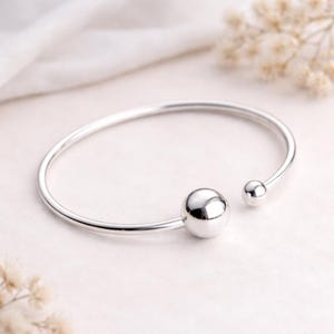 Könnte beinhalten: Ein offenes, silberfarbenes Armreif-Armband mit zwei kugelförmigen Akzenten. Die größere Kugel befindet sich in der Mitte, die kleinere am offenen Ende. Das Armband liegt auf einer weißen Oberfläche.