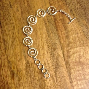 Puede incluir: Pulsera plateada con una serie de diseños en espiral conectados por pequeños eslabones. La pulsera tiene un cierre de palanca. La joya se exhibe sobre una superficie de madera.