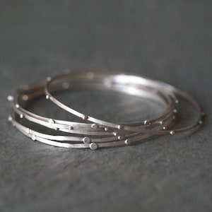 May include: A collection of silver bangles with a textured, dotted pattern. The bangles are slender and circular, featuring small, evenly spaced dots along their surface. The bracelets are stacked together, creating a layered effect. The background is a muted grey.